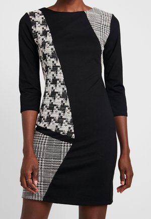 Schwarzes Kleid mit dreiviertel Ärmeln, vorne und an der linken Hüfte mit schwarz-weißen Hahnentritt- und Karomustern in geometrischem Design.