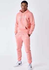 Rosa Kapuzenpullover und passende Jogginghosen, beide aus weichem Stoff, mit weißem Textlogo. Schmale Passform mit gerippten Bündchen und Bund.