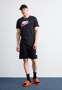Muž v černém tričku Nike, černých šortkách Nike, černé kšiltovce, černých ponožkách a oranžových pantoflích stojí na modrém koberci proti bílé zdi.