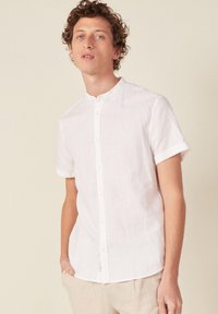 Camicia bianca in lino a maniche corte con colletto alla coreana e chiusura con bottoni; presenta polsi arrotolati e un tessuto leggero e strutturato.