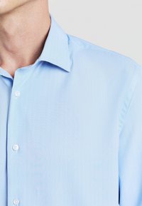 Camicia button-up azzurro chiaro con un tessuto testurizzato, colletto a punta e bottoni bianchi, inquadratura ravvicinata della parte superiore del corpo.