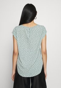 Blusa verde con lunares, mangas cortas, tejido suave, cuello redondo y cierre de cremallera en la parte trasera. Presenta un dobladillo curvo y una textura sutil.