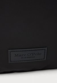 Černá, texturovaná látková taška s obdélníkovou embosovanou nášivkou loga s nápisem "Marc O'Polo" a "EST. IN STOCKHOLM" v tmavém písmu.