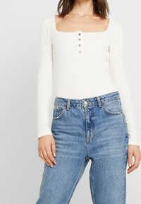 Body a maniche lunghe bianco a coste con scollatura quadrata e sei bottoni a pressione metallici, abbinato a jeans blu a vita alta.