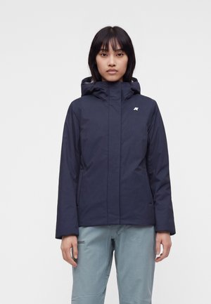 Femme debout portant une veste à capuche bleu marine et un pantalon bleu clair sur un fond clair uni.