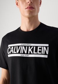 Young man wearing black T-shirt with large white Calvin Klein New York logo on the chest against a plain background.