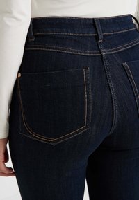 Jean en denim bleu foncé avec des coutures orange contrastantes, comprenant deux poches arrière et une taille haute. Texture lisse.