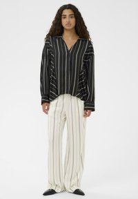 Ženska z dolgimi kodri, oblečena v črno-belo verticalno prugasto srajco in bele široke hlače s črnimi prugami, obuta v črne čevlje.