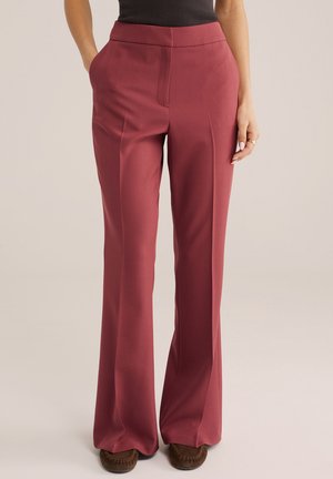 Donna che indossa pantaloni a gamba larga a vita alta di colore rosa con tasche laterali e mocassini marroni, in piedi contro uno sfondo neutro.