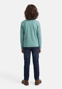 Παιδί με καστανά σγουρά μαλλιά στέκεται βλέποντας προς τα πίσω, φορώντας μπλε-τεϊρκουάζ μακρυμάνικο πουκάμισο, σκούρο μπλε τζιν και καστανά παπούτσια σε λευκό φόντο.