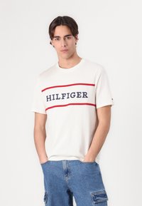 White t-shirt with a crew neck, featuring "HILFIGER" in navy text, bordered by red lines. Soft fabric, paired with blue denim cargo shorts.