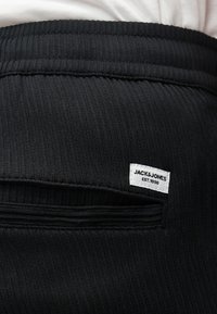 Μαύρο ριμπ ύφασμα παντελόνι με τσέπη που φέρει μια λευκή ετικέτα με την επιγραφή "JACK & JONES EST. 1990" κοντά στη μέση.