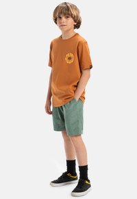 Bruine katoenen t-shirt met een gele ronde grafische print, gecombineerd met groene corduroy shorts en zwarte sneakers met gele accenten.