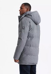 Parka imbottita grigia con cappuccio, dotata di una chiusura frontale con bottoni e tasche laterali. Include una patch quadrata con logo sul braccio sinistro.