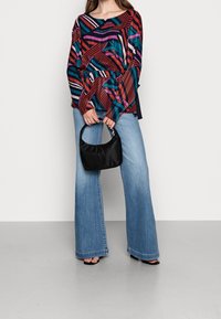 Haut à manches longues coloré avec des motifs abstraits, associé à un jean en denim à jambes larges et un petit sac à main noir, sur un fond de sol gris.