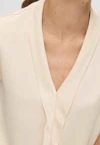 Krémová blůza s výstřihem do V. Vyrobená z hladké tkaniny, s jemným drapováním a minimálními švy na límci.