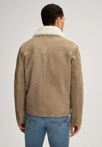 Giacca in suede marrone con colletto in shearling bianco, caratterizzata da un design leggermente aderente e cucitura a yoke sul retro. Indossata con jeans blu.