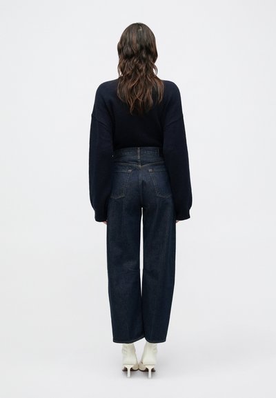 Tengeri kék pulóver túlméretezett ujjakkal, magas derekú sötét farmer széles szárú nadrággal és fehér, sarkú bokacsizmával párosítva.