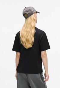 Juodas medvilnės marškinėliai su trumpomis rankovėmis, dėvimi su pilkomis treniruočių kelnėmis. Temoje yra ilgi, bangoti šviesūs plaukai ir pilkas kepuraitė.