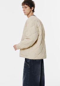 Beige kviltet jakke med afslappet pasform, rund hals og elastiske manchetter, parret med mørke denimbukser. Glat tekstur.