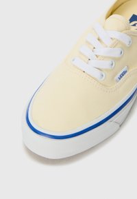 Yellow canvas sneaker with white laces, blue accents on the sole, and logo tag, featuring a rounded toe and textured surface.