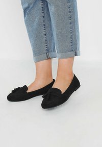 Svarta mockaloafers med en bård detalj på framsidan, platt sula och mjuk yta, tillsammans med uppvikta ljusa denimjeans.