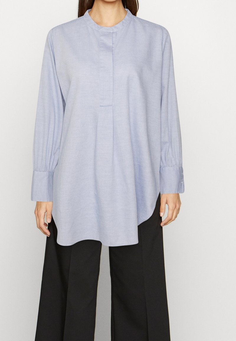 Femme portant une blouse tunique bleu clair à manches longues avec col officier et pantalon noir à jambes larges, debout sur un fond blanc.