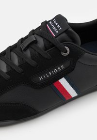 Baskets noires Tommy Hilfiger avec lacets noirs, logo sur la languette et le côté, avec un détail en tissu rayé rouge, blanc et bleu.
