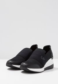 Scarpe nere senza lacci con parte superiore in rete e cinghie elastiche. Presentano una suola bianca rialzata, accenti testurizzati e un dettaglio del logo sul lato.