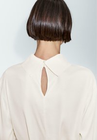Blouse de couleur crème avec un col et une ouverture en forme de clé dans le dos, au toucher lisse et à la coupe fluide.