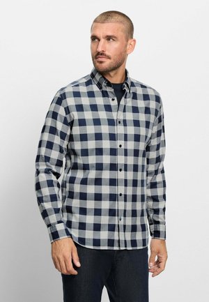 Mann mit kurzen Haaren und Bart, trägt ein grau-navyblau kariertes Hemd mit Knöpfen und dunkle Hose, schaut nach links.