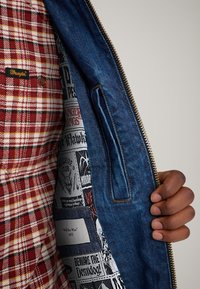 Red and yellow plaid flannel interior with brown buttons, denim exterior with a gold zipper, featuring patterned fabric lining and a pocket.