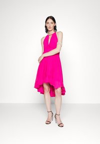 Abito rosa plissettato con collo a halter, orlo asimmetrico e dettagli a volants. La modella indossa tacchi neri con cinturini e un bracciale d'argento.