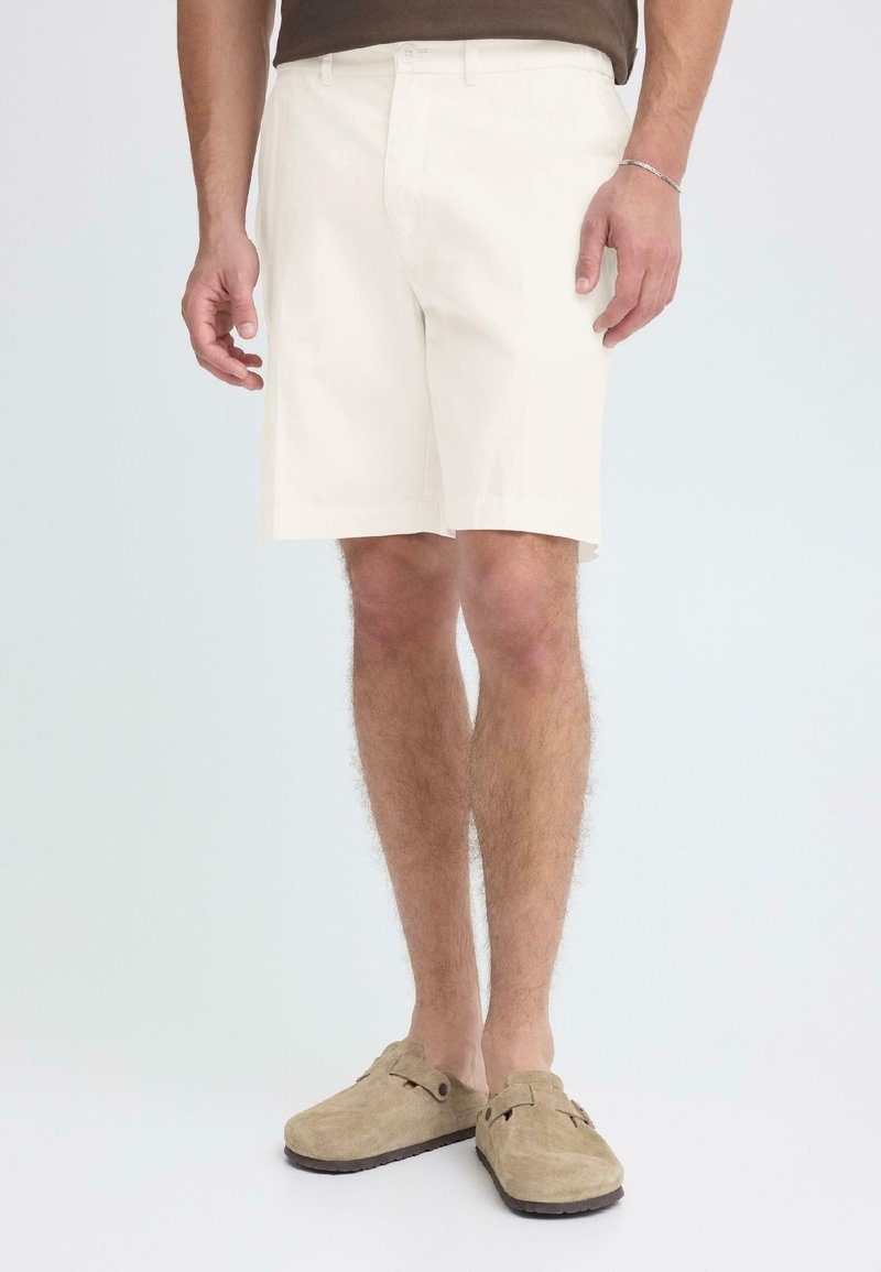 Shorts de couleur claire en tissu à texture lisse. Dotés de poches standard et d'une coupe droite ; portés avec des chaussures beiges sans lacets.