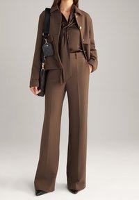 Femme portant un pantalon marron sur mesure, une veste assortie avec des boutons dorés, un chemisier en soie et un sac à main noir porté à l'épaule.