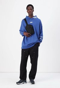 Modrá oversized mikina, čierne nohavice so striebornými akcentmi a čierna crossbody taška. Model má na sebe šedé tenisky a stojí na jednoduchej časti pozadia.
