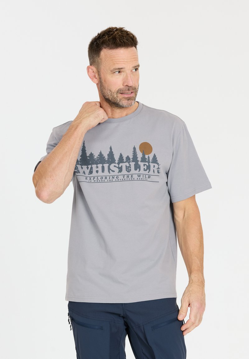 Mann mit kurzem Haar und Bart, der ein hellgraues "Whistler"-T-Shirt mit Wald- und Sonnenmotiv trägt, schaut zur Seite und berührt dabei seinen Hals.