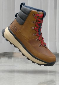 Botte de randonnée en cuir marron avec un col rembourré gris, des lacets rouges, une semelle en caoutchouc noire et un logo de marque bien visible sur le côté.