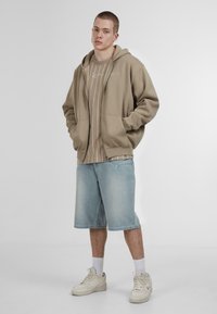 Beige zip-up hoodie over a light striped shirt, paired with light blue denim shorts and white sneakers, featuring subtle patterns and textures.