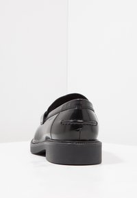 Sapato de verniz preto com um salto baixo, forma arredondada e superfície lisa e brilhante. Apresenta costura mínima e um design de calçar fácil.