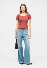 Γυναίκα με κυματιστά μαλλιά που φοράει τοπ με κοντά μανίκια σε σκουριά απόχρωση, ανοιχτό μπλε τζιν παντελόνι με φαρδιά μπατζάκια, μαύρα παπούτσια και κρατάει μαύρη τσάντα.