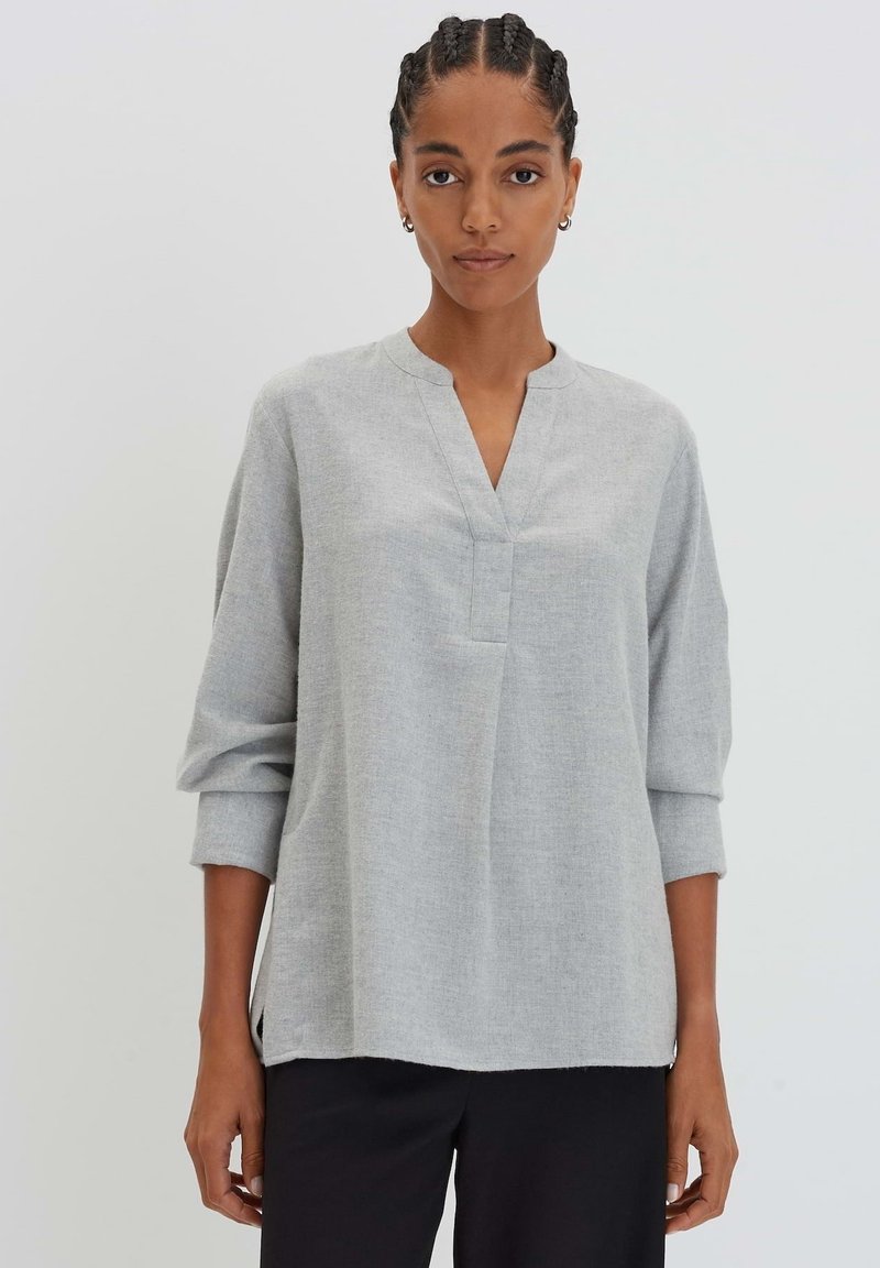 Tunique gris clair avec coupe décontractée, décolleté en V profond, manches trois-quarts et fentes sur les côtés. Texture douce et détails de couture minimaux.