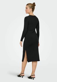Abito nero a coste con maniche lunghe, scollatura rotonda e spacco laterale, realizzato in tessuto elasticizzato, abbinato a sandali neri con punta aperta.