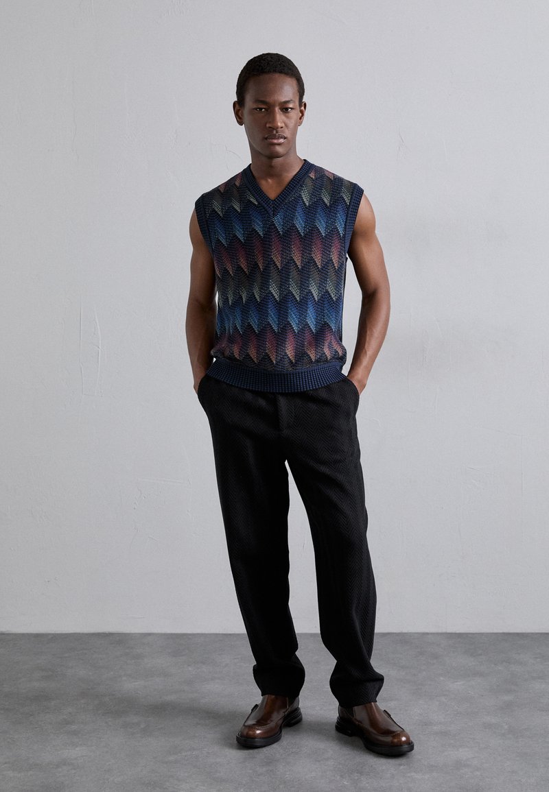 Maglione a maniche corte con scollatura a V, caratterizzato da un motivo a chevron testurizzato nei toni del blu, rosso e giallo, abbinato a pantaloni a scacchi houndstooth neri e scarpe marroni.