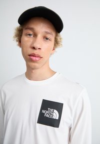 Weißes Langarmshirt mit einer schwarzen quadratischen Tasche, die weißen Text und ein Logo zeigt. Eine schwarze Kappe rundet den lässigen Look ab. Glatter Stofftextur.