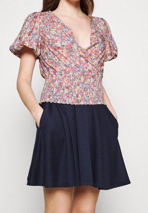 Vrouw die een bloemrijk roze, blauw en geel topje met pofmouwen en een gerimpelde taille draagt, gecombineerd met een donkerblauwe A-lijn rok, handen in de zakken.