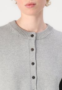 Hellgrauer Strickpullover mit einem gerippten Kragen, ausgestattet mit fünf dunklen Knöpfen und einer glatten Textur. Einfaches Design, ungeschmückte Vorderseite.