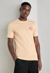 Beige T-Shirt mit kurzen Ärmeln aus weichem Stoff, mit einem kleinen weinroten Grafikdesign auf der Brust. Rundhalsausschnitt und slim-fit Silhouette.