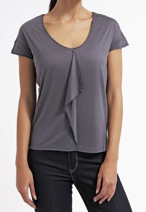 Vrouw draagt een grijze top met korte mouwen, een V-hals en een centrale ruches, gecombineerd met een donkere spijkerbroek tegen een witte achtergrond.