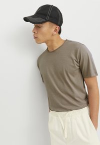 Camiseta marrón de manga corta con cuello redondo, acompañada de pantalones de color crema con cordón, y una gorra negra con costuras en contraste.